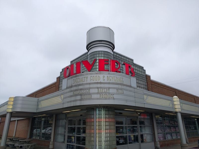 Art-Deco style store exterior with curved, backlit glass and etched signage that reads "Specialty Food and Beverages, Delicatessen, Bakery, Pizza, Liquor, Produce