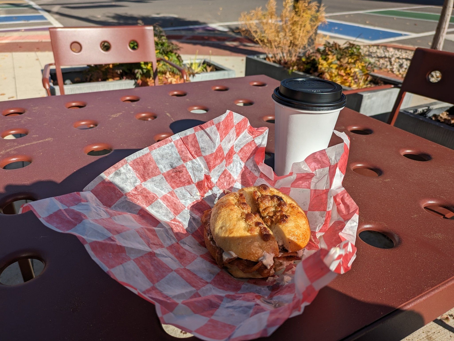 French bagel with bacon and maple brown sugar and a chai tea in a to-go wrapper on a table inside an outdoor seating area along the sidewalk