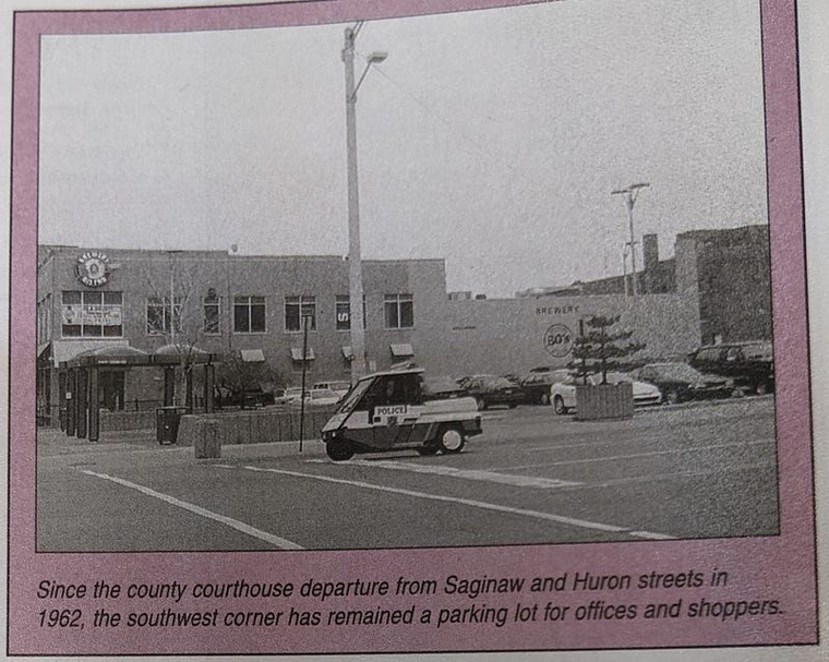 A photo of a page from a book showing a small police golf cart passing the shelters. The printed caption reads: "Since the county courthouse departure from Saginaw and Huron streets in 1962, the southwest corner has remained a parking lot for offices and shoppers."