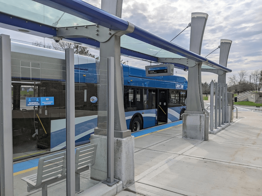 A blue and white articulated bus at a level boarding platform with its doors open. The platform has a shelter, a departure display, and several seats.