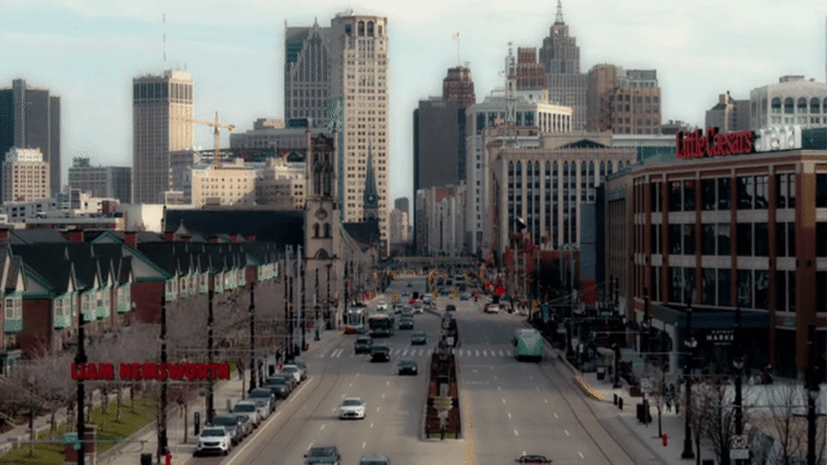 An aerial shot of Woodward Avenuefrom north of Little Caesars Arena, looking south toward downtown. Two DDOT buses and the QLINE streetcar are visible