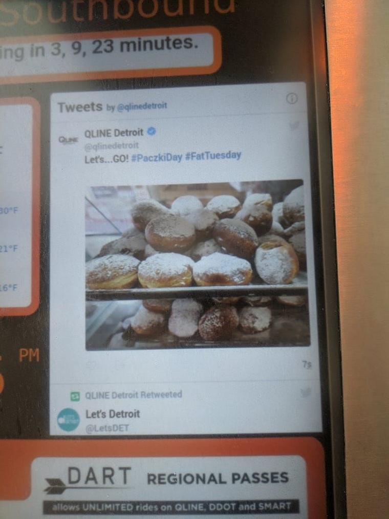 The QLINE departure board showing the agency's latest Tweet, a picture of paczki with caption "Let's... GO! #PaczkiDay #FatTuesday"