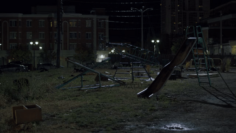 Broken playground equipment in an overgrown field between old buildings