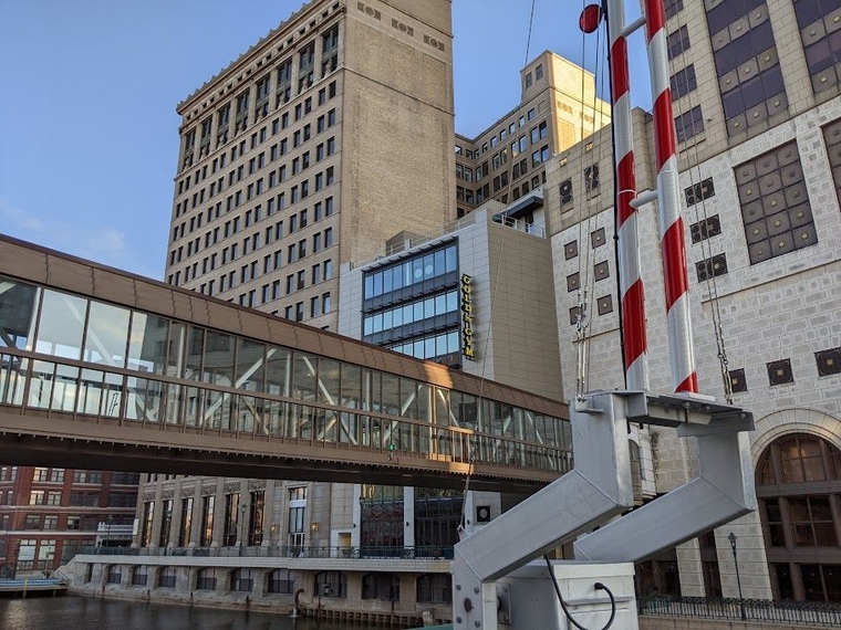 A covered walkway over an inlet to the river, with a raised drawbridge arm visible in the foreground and several brick and concrete buildings in the background.