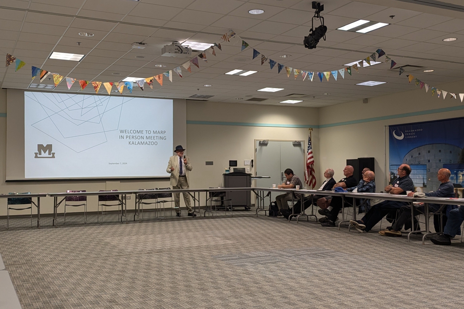 Mark Buckley, in a tan suit and black top hat, addresses meeting attendees in front of a projector slide reading "Welcome to MARP In-Person Meeting, Kalamazoo".