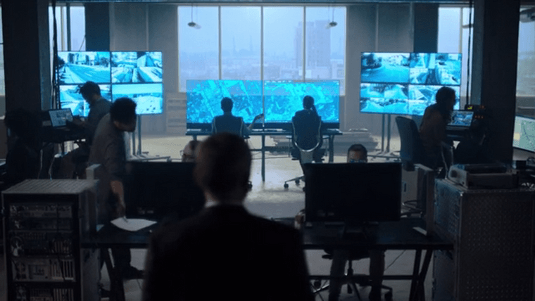 Miles’s control room, featuring multiple large monitors with maps and CCTV footage in shades of blue