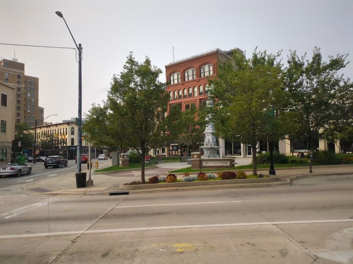 A brown brick building behind a plaza with trees and a statue as seen from the bus window