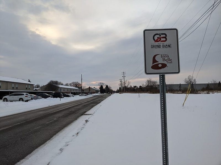 A signpole reads "City of Grand Blanc: Iron Belle Trail" on Reid Road on a snowy day.