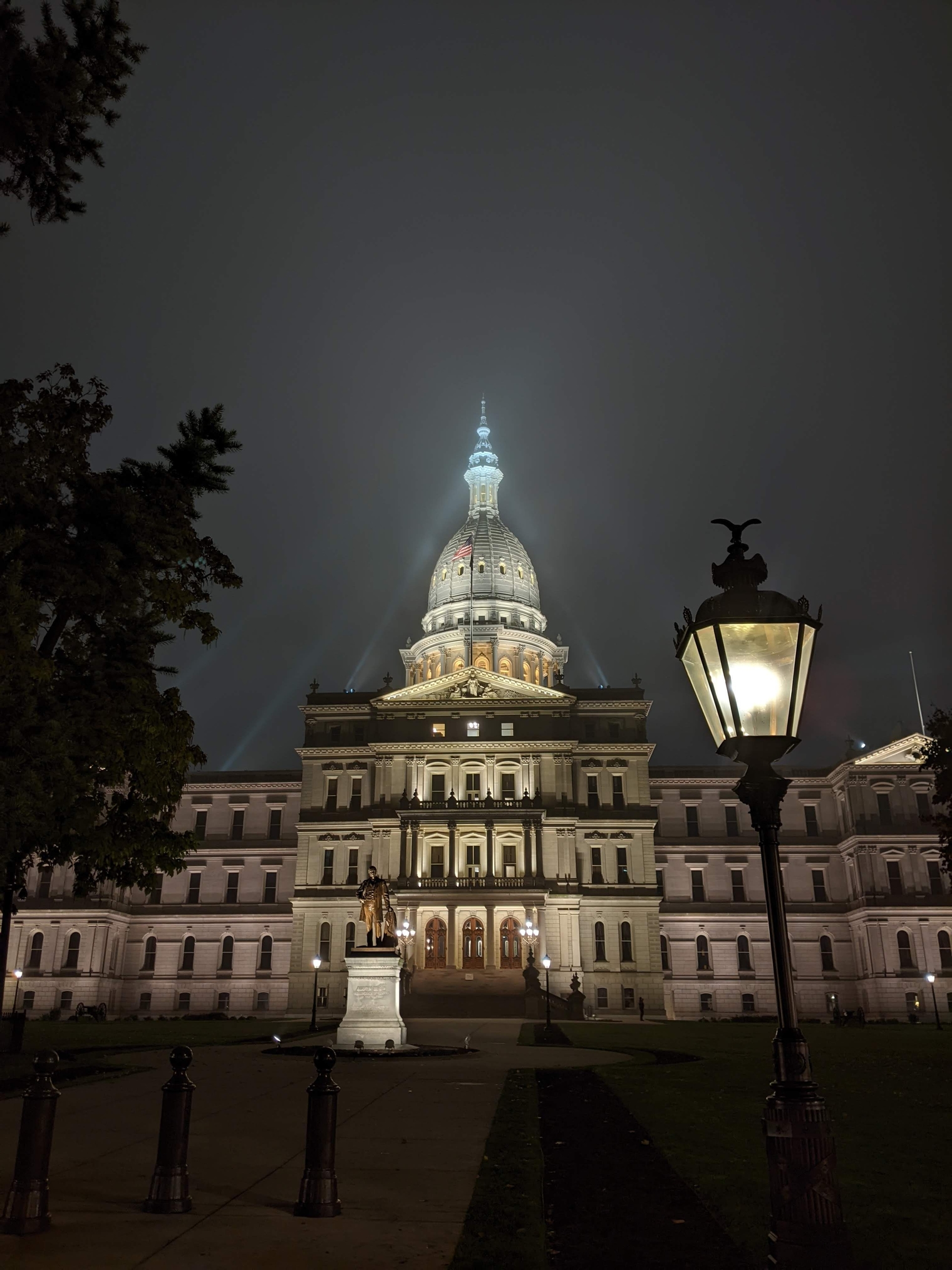 The Michigan State Capitol on a foggy night with the dome illuminated by multiple floodlights. A lamp along the east lawn pathway shines through the fog in the foreground.