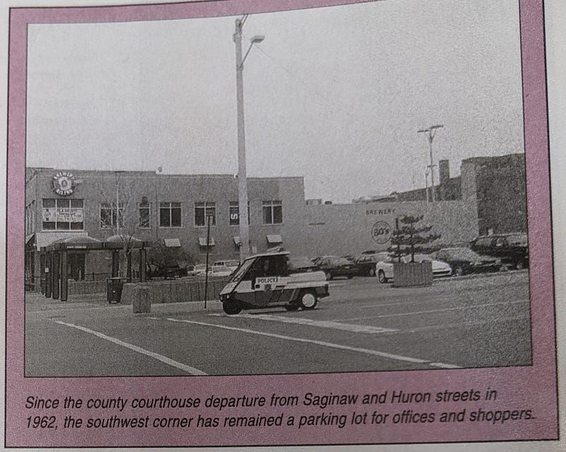 A photo of a page from a book showing a small police golf cart passing the shelters. The printed caption reads: "Since the county courthouse departure from Saginaw and Huron streets in 1962, the southwest corner has remained a parking lot for offices and shoppers."