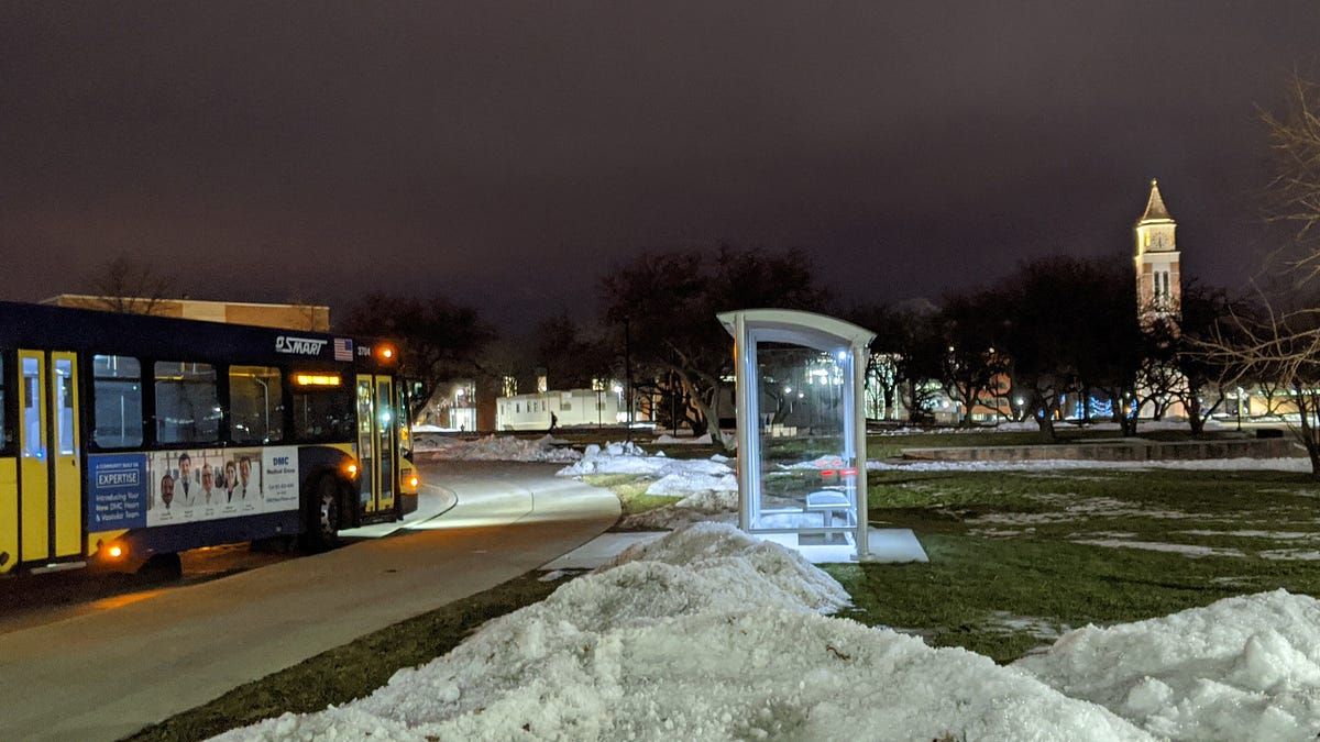 A blue and yellow SMART bus approaches a shelter at night with Oakland University’s clock tower in the background