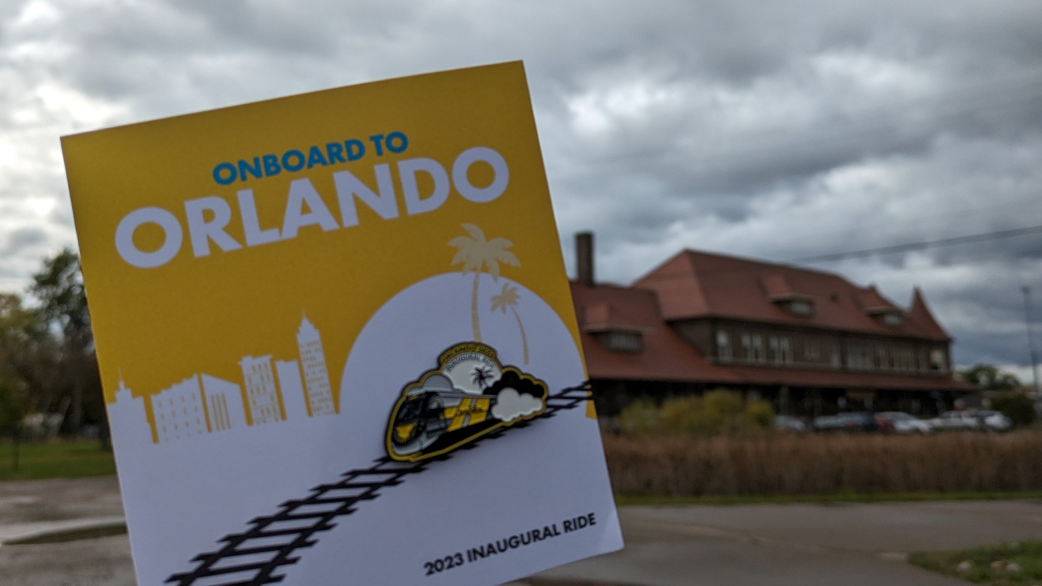 Me holding up the pin, a small caricature of a Brightline train, outside Durand Station