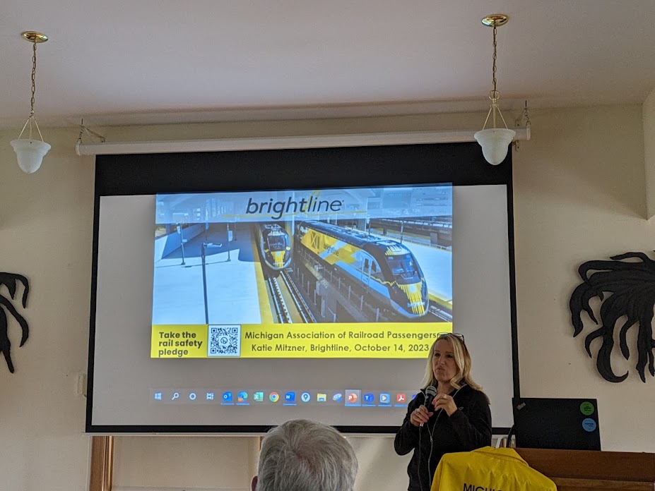 Katie Mitzner holds a microphone while addressing a large audience from beside a lectern. A slide on a projector screen shows a picture of two Brightline trains at a station platform in Florida.