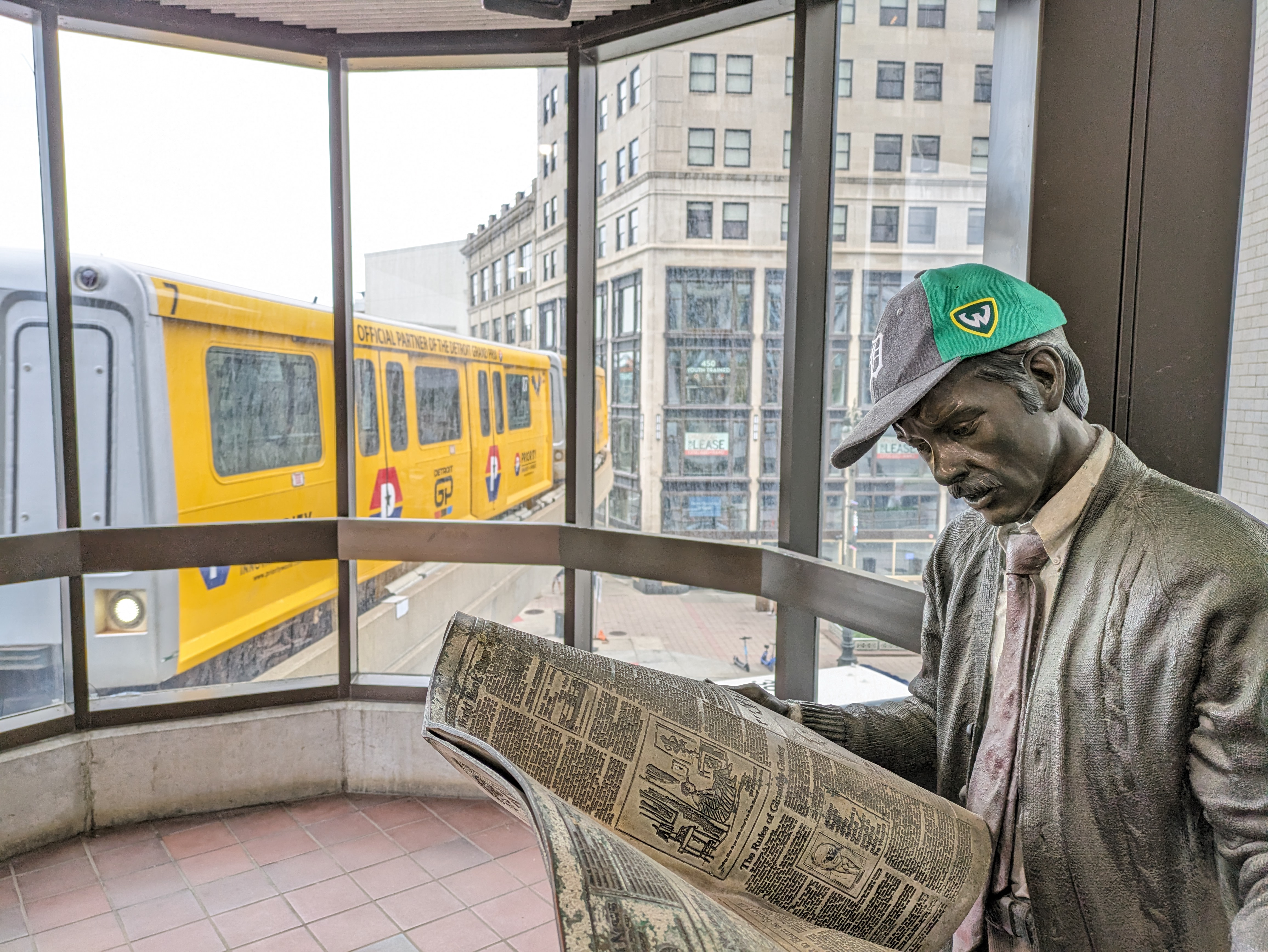 My Tigers Wayne State hat on the "Catching Up" statue of a man reading the Detroit News on the Grand Circus Park People Mover platform