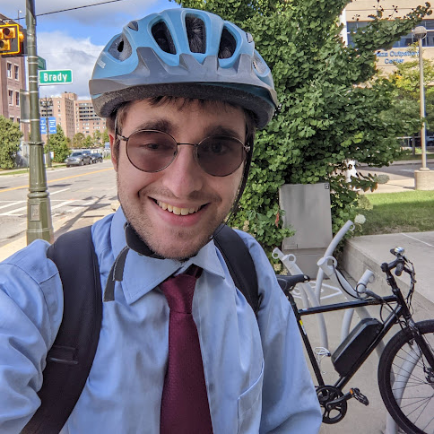 Corey in a blue dress shirt and red tie wearing a helmet with his bike in the background
