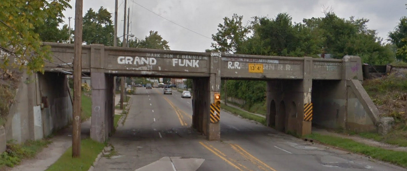 A short railroad bridge with a flashing warning light to signal low clearance. "Grand Funk" has been painted over the original markings which read "Grand Trunk Western"