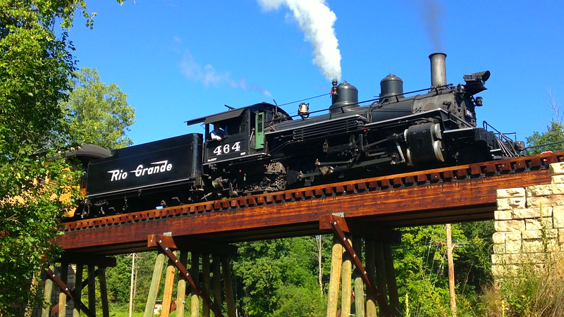 A black historic steam locomotive, Rio Grande Engine 464, crosses the brown wooden Butternut Creek Trestle at Crossroads Village in Flint.